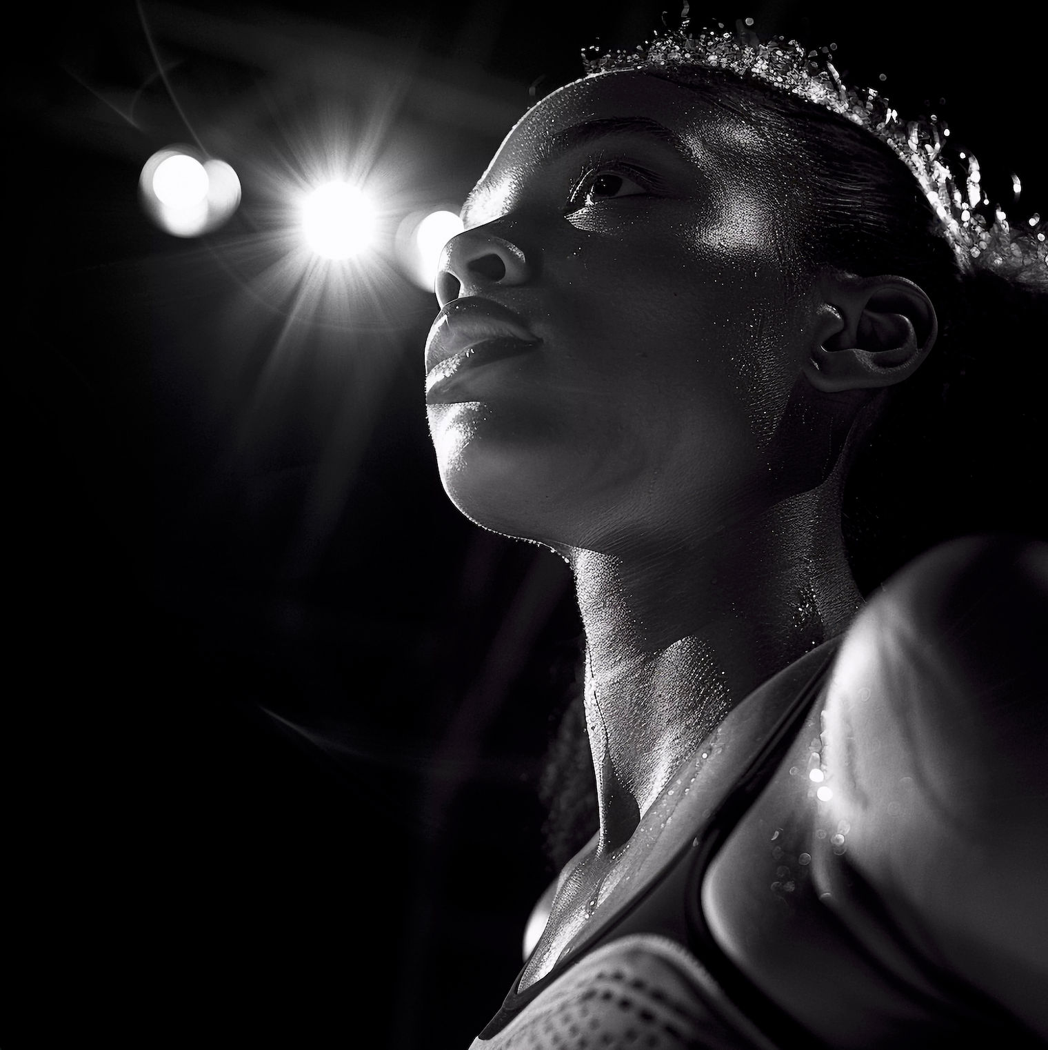A dramatic black and white low-angle shot of a sweaty woman in athletic wear looking upward, with bright stadium-style lights creating a lens flare behind her.
