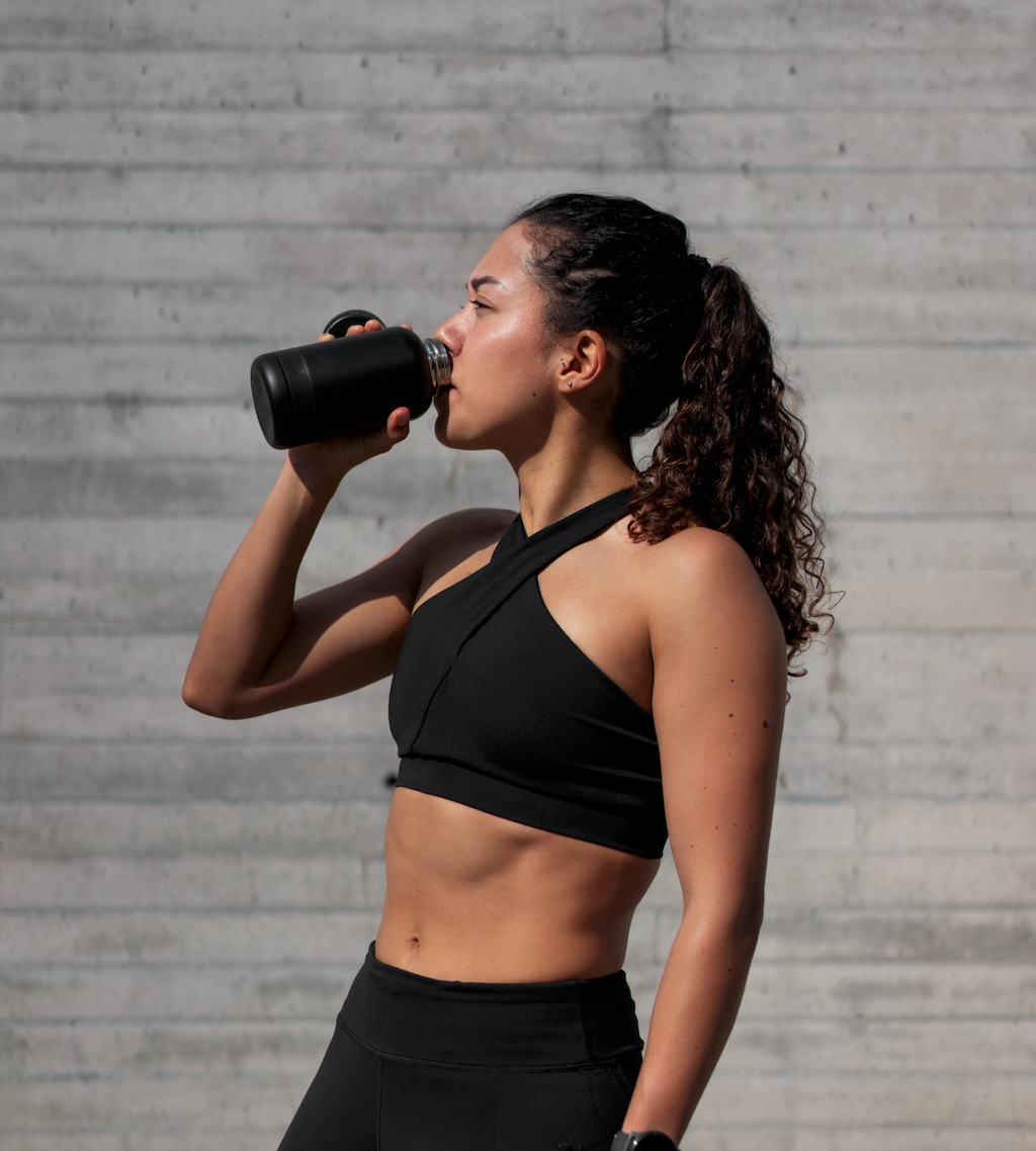 A side profile of a woman with curly hair in a ponytail, wearing a black cross-neck sports bra and leggings, as she drinks from a black water bottle against a grey concrete wall in bright sunlight.
