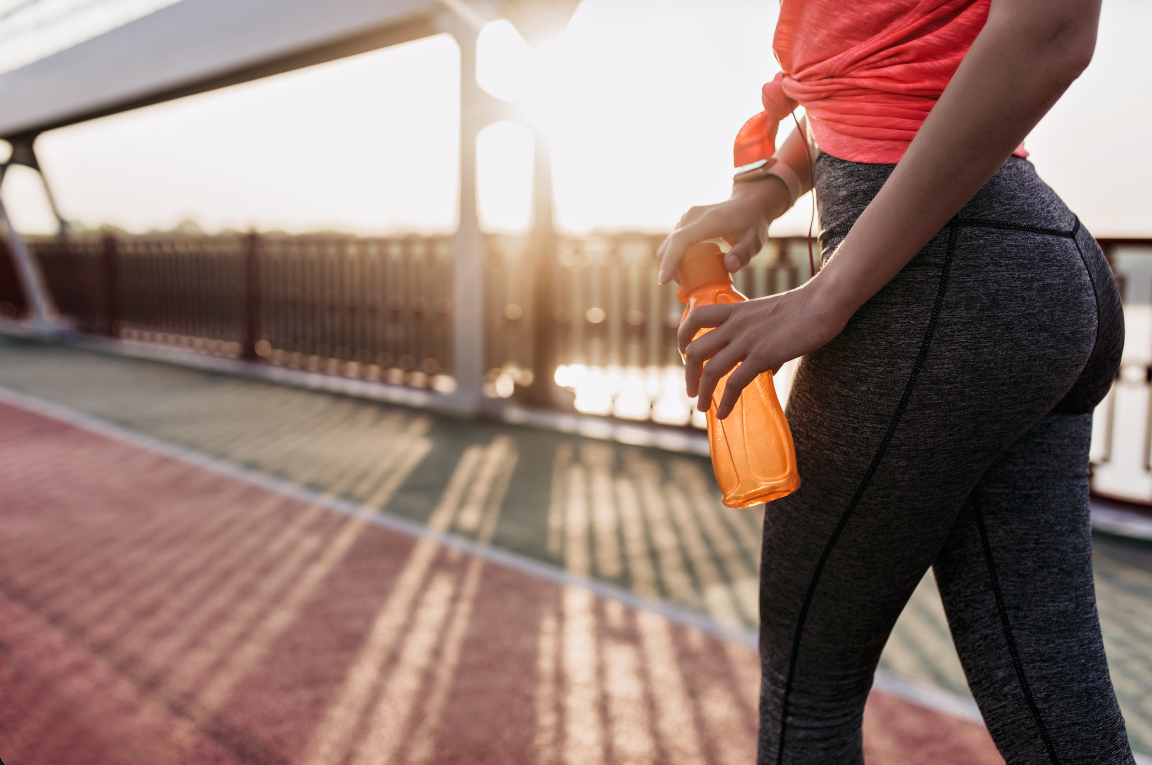 A lower-body shot of a woman in grey leggings and an orange athletic top holding an orange water bottle while walking on a bridge at sunset.