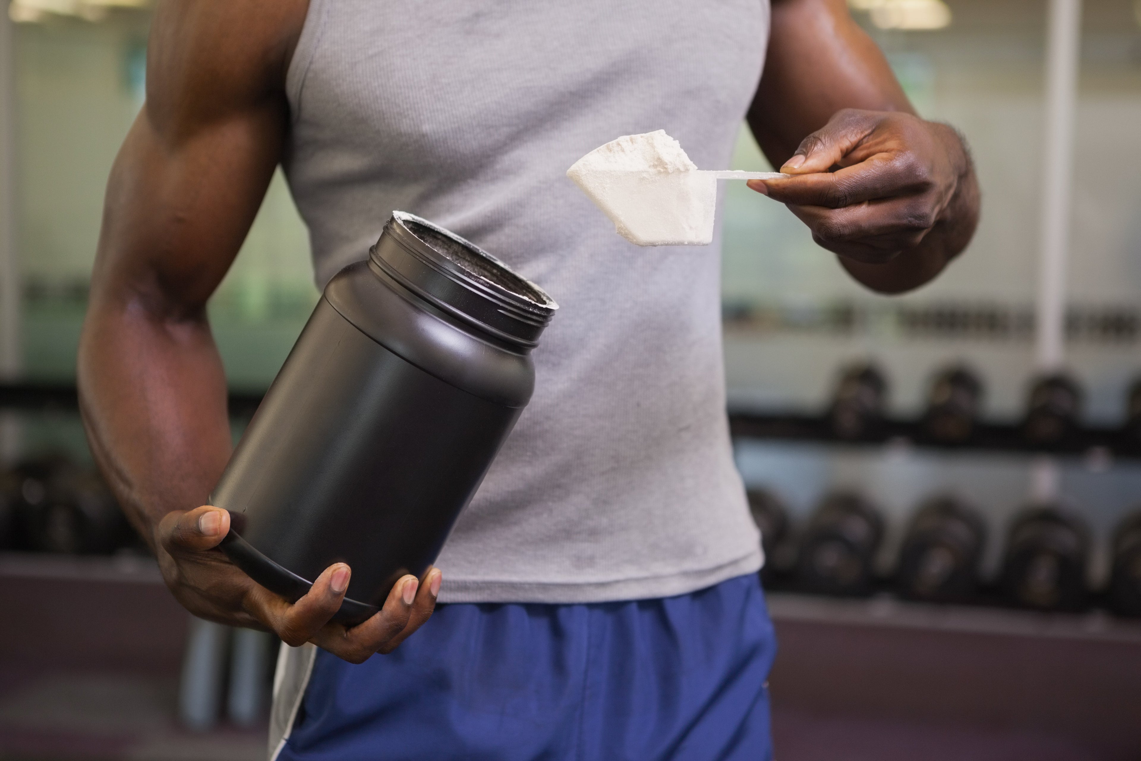 A man in a grey tank top and blue shorts holds a large black supplement tub and a scoop of white powder in a gym setting with a dumbbell rack in the background.