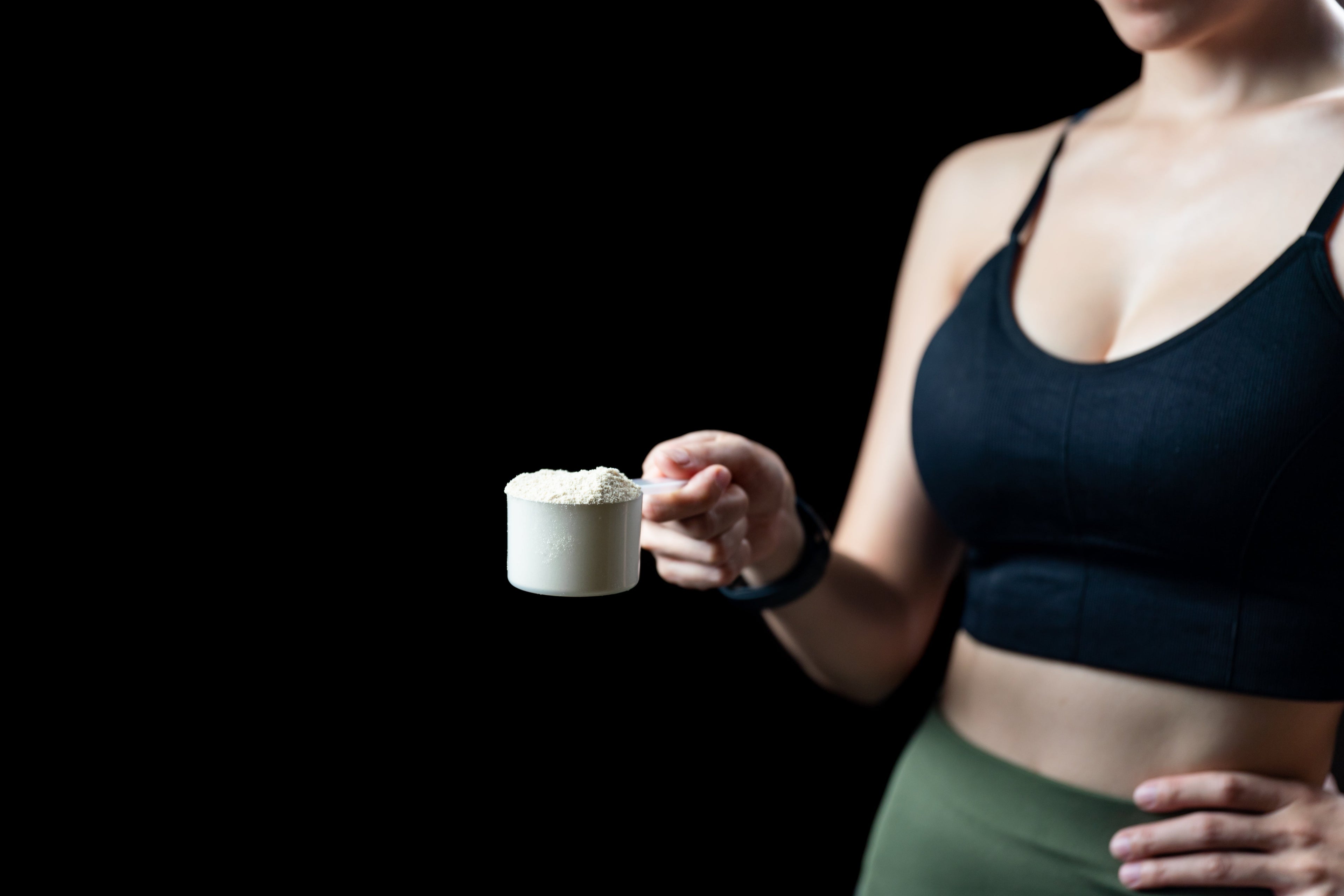 A woman wearing a black sports bra and green leggings holds a white scoop filled with supplement powder out toward the camera against a solid black background.