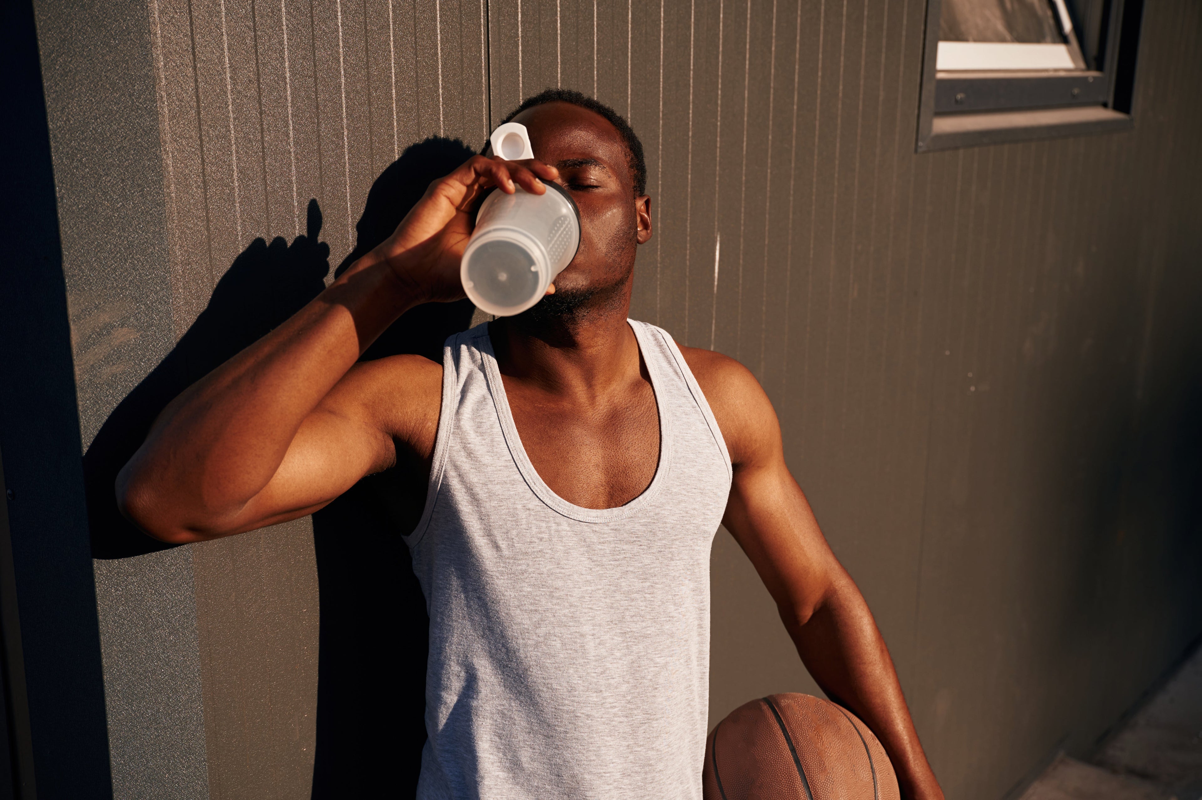 A man in a grey tank top leans against a dark, textured wall outdoors while drinking from a shaker bottle. He is holding a basketball under his arm, and the scene is lit by warm, low-angle sunlight.