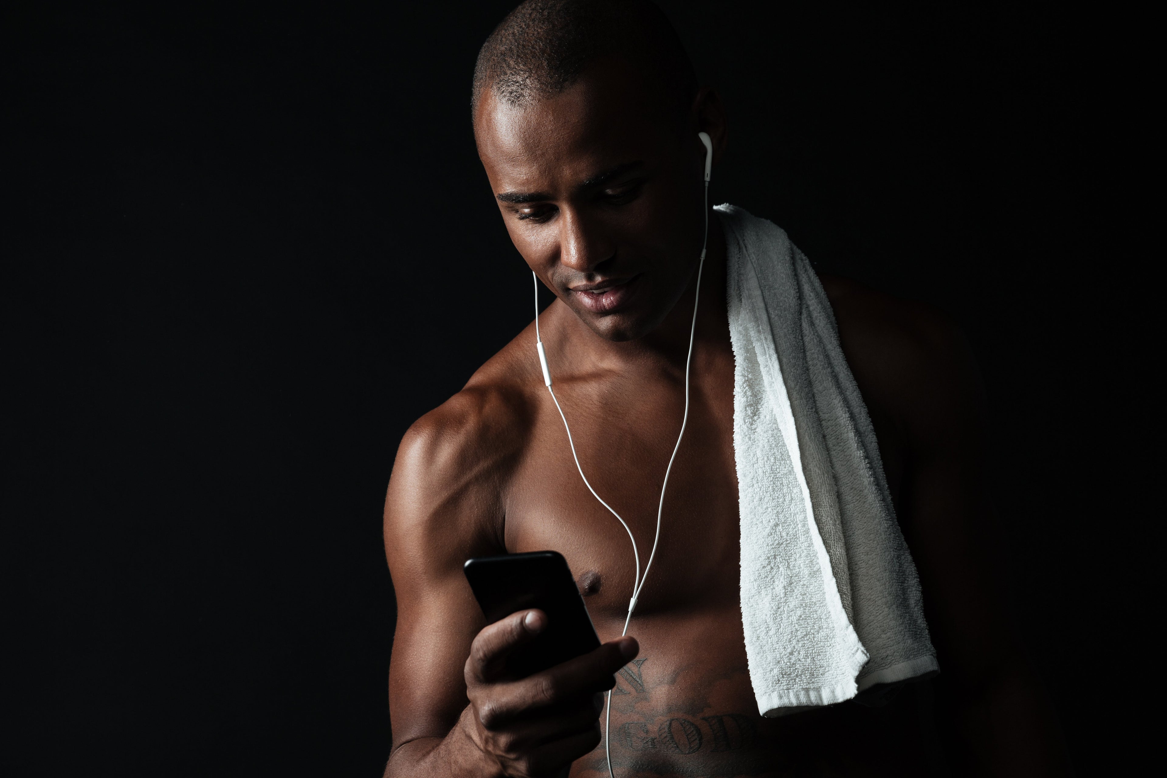 A shirtless man with a white towel draped over his shoulder and white wired earbuds looks down at his smartphone against a dark background.