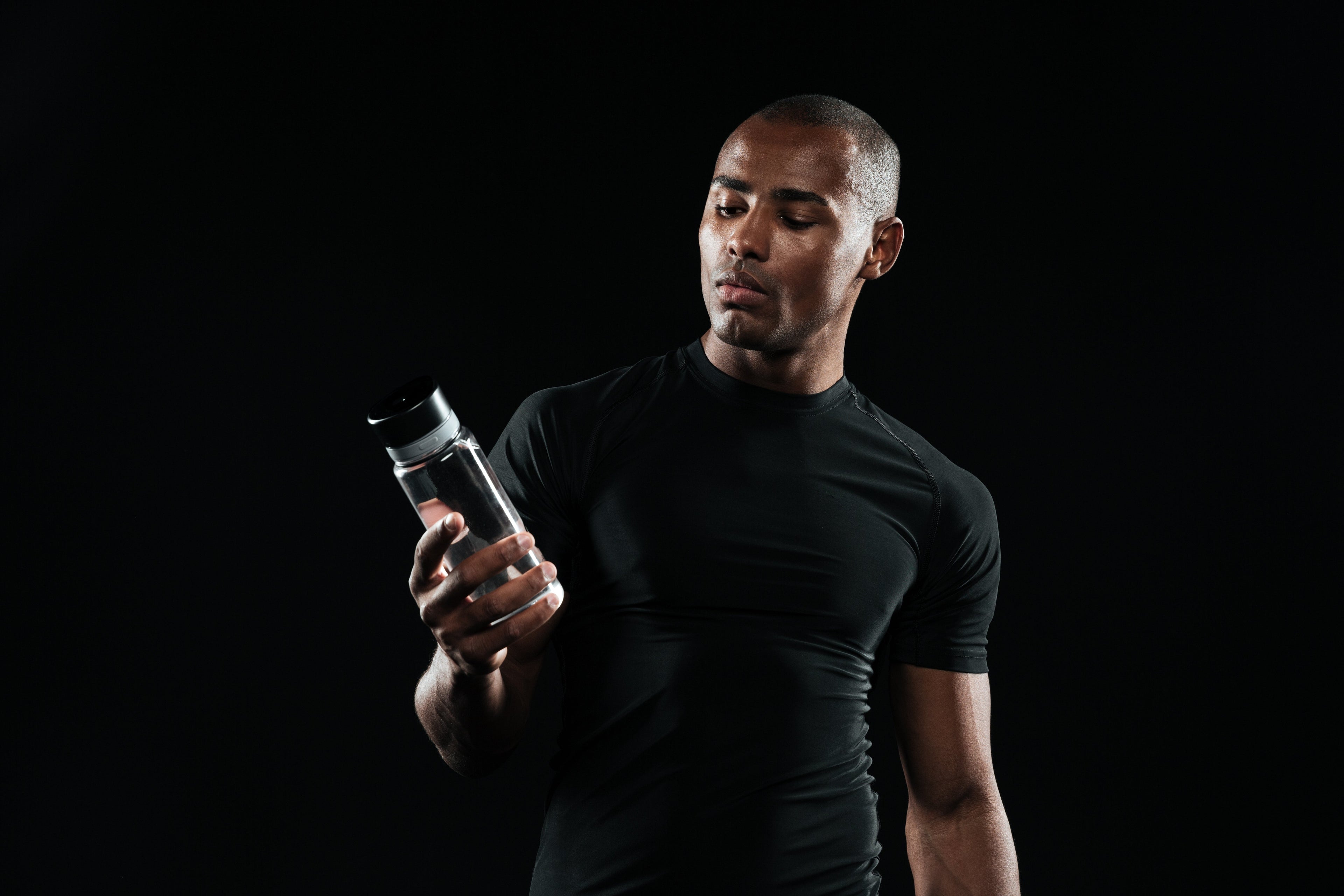 A man in a tight black athletic shirt stands against a black background, looking intently at a clear water bottle he is holding in his hand.