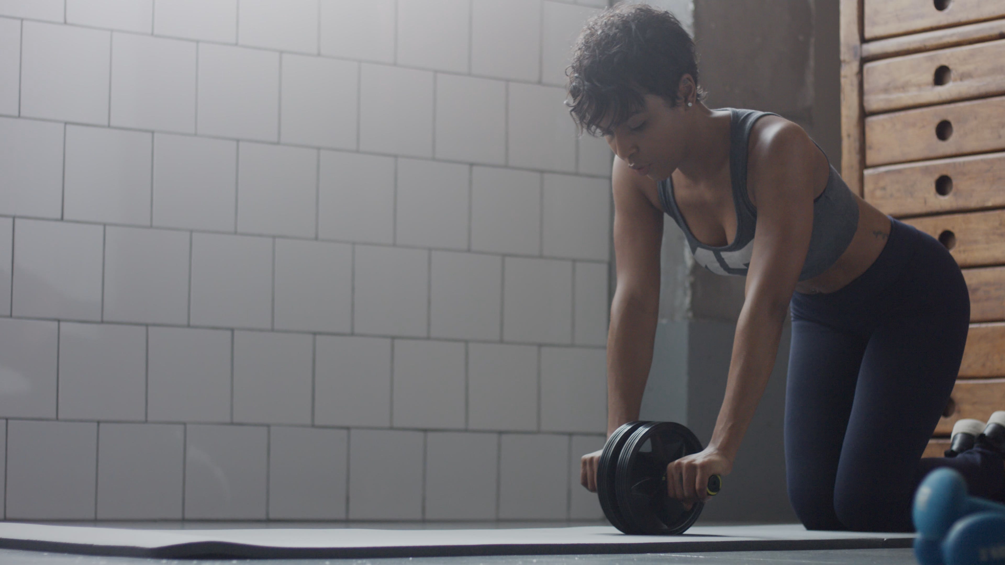 A woman in a grey sports bra and dark leggings performs an ab-rollout exercise using an ab wheel on a mat in a gym with white tiled walls.
